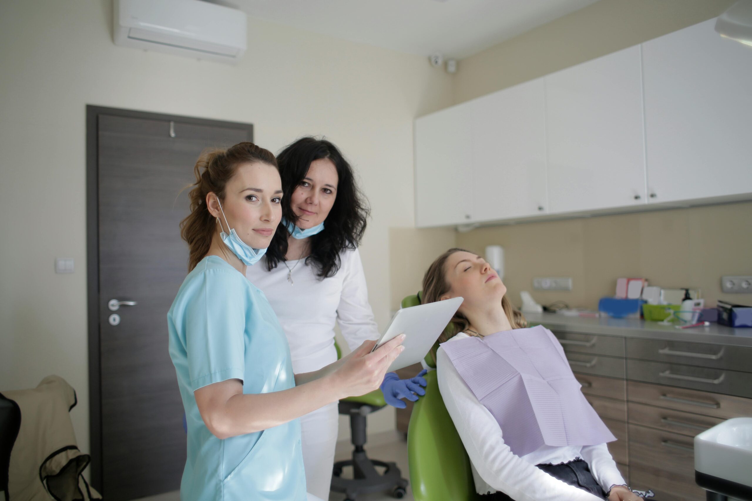 Smiling dentist in sterile mask and uniform looking at camera and showing tablet screen to cheerful female assistant while standing near patient with closed eyes sitting on chair in clinic